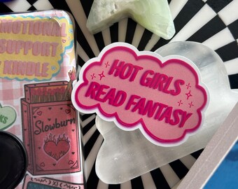 Könnte beinhalten: Ein rosa-weißer Aufkleber in Wolkenform mit dem Text "Hot Girls Read Fantasy". Der Aufkleber befindet sich auf einer weißen Oberfläche mit einem schwarz-weißen Schachbrettmuster.