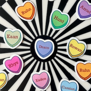 May include: Heart-shaped stickers with names like "Rhys", "Rowan", and "Draco" are scattered on a black and white target design. Each sticker has a different color, including pink, yellow, and green, with a contrasting border and text.