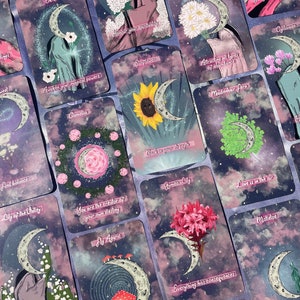 Puede incluir: Un conjunto de 16 cartas de tarot con una luna creciente y varias flores y plantas. Las cartas tienen un fondo morado y rosa con un efecto de cielo estrellado. Las cartas presentan texto como "Time to rest", "Okra", "Honour your spirituality", "Roman Chamomile", "You glow with confidence", "Awaken your magical powers", "Sunflower", "Cyclamen", "Attracting all types of abundance", "Camellia", "Maidenhair fern", "Lily of the Valley", "With pain comes growth", "Back in your strength", "You are the creator of your own destiny", "Gymea Lily", "Love is in the air", "Fly Agaric", "Everything has consequences", "Mistletoe", "Buttercup", "Beware of deceitful toxicity", and "Defying".