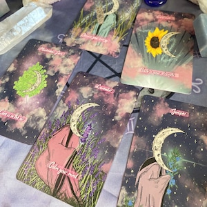 Puede incluir: Un juego de cartas de tarot con un tema celestial. Cada carta presenta una planta diferente y una luna creciente. Las cartas tienen un esquema de colores rosa, morado y azul con un fondo estrellado. El texto en las cartas incluye "El amor est&aacute; en el aire", "Helecho maidenhair", "Limpia tu energ&iacute;a", "Lavanda", "Calma tu mente", "De vuelta a tu fuerza", "Enebro" y "Det&eacute;n la propagaci&oacute;n".