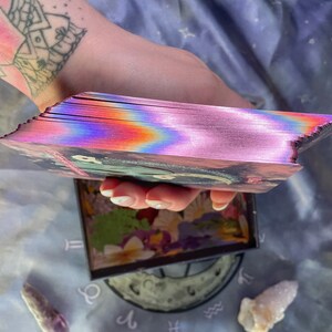 Puede incluir: Una baraja de cartas de tarot con un dise&ntilde;o hologr&aacute;fico, iridiscente, rosa, azul y morado. Las cartas est&aacute;n apiladas y desplegadas, mostrando los bordes de las cartas.