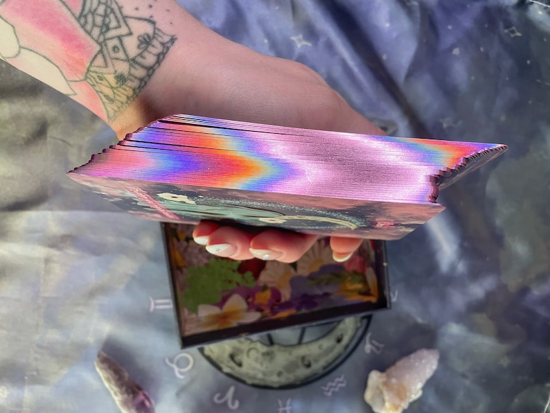 Puede incluir: Una baraja de cartas de tarot con un dise&ntilde;o hologr&aacute;fico, iridiscente, rosa, azul y morado. Las cartas est&aacute;n apiladas y desplegadas, mostrando los bordes de las cartas.