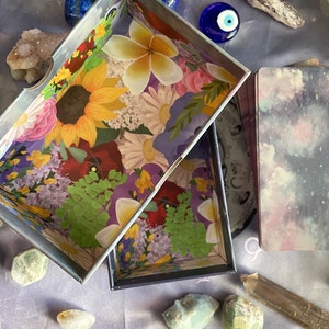 Puede incluir: Dos cajas abiertas con dise&ntilde;os florales y gal&aacute;cticos. La caja floral tiene un patr&oacute;n floral colorido con un girasol, flores rojas y hojas verdes. La caja de galaxia tiene un fondo azul oscuro con nubes blancas y estrellas. Las cajas est&aacute;n sobre una superficie morada con cristales y otros art&iacute;culos decorativos.