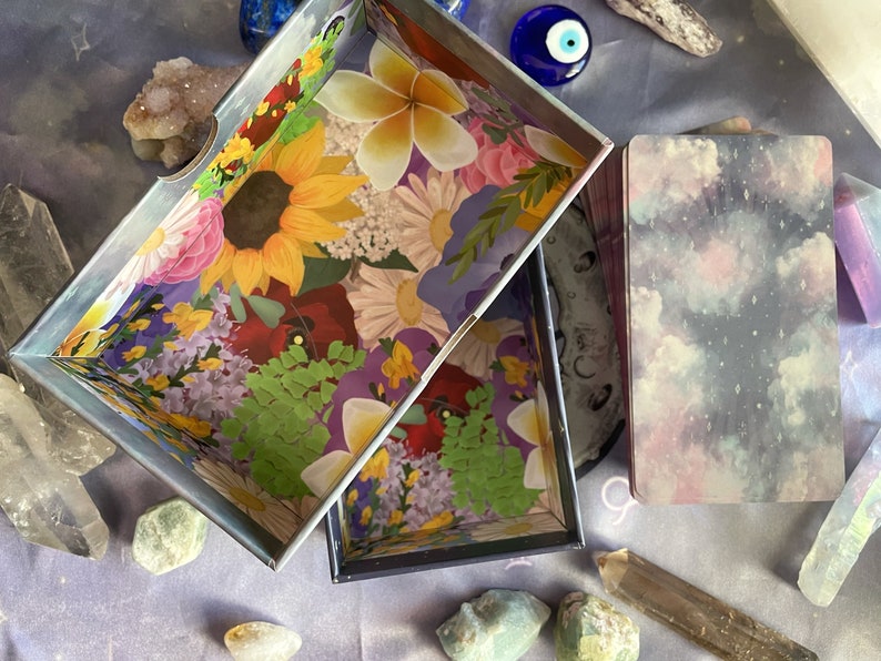Puede incluir: Dos cajas abiertas con dise&ntilde;os florales y gal&aacute;cticos. La caja floral tiene un patr&oacute;n floral colorido con un girasol, flores rojas y hojas verdes. La caja de galaxia tiene un fondo azul oscuro con nubes blancas y estrellas. Las cajas est&aacute;n sobre una superficie morada con cristales y otros art&iacute;culos decorativos.