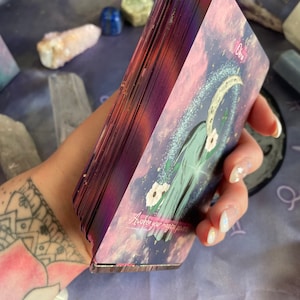 Puede incluir: Una baraja de cartas de tarot con un fondo morado y rosa. La carta en primer plano muestra una luna creciente y el texto "Awaken your magic power".