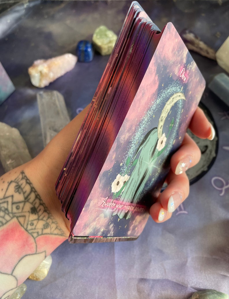 Puede incluir: Una baraja de cartas de tarot con un fondo morado y rosa. La carta en primer plano muestra una luna creciente y el texto "Awaken your magic power".