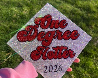 May include: A graduation cap with a sparkly, silver surface. The words "One Degree Hotter" are written in red rhinestones, with the year "2026" below. The cap is held up against a background of green grass.