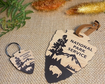 May include: Wooden National Park Service keychain and luggage tag. The tag has a leather strap. Both items feature a black silhouette of a tree, mountains, and a bison, with the words "National Park Service".