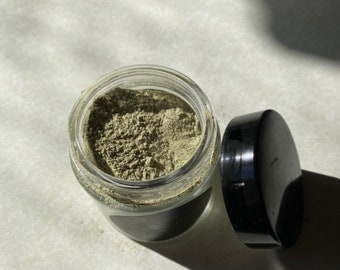 May include: A glass jar with a black lid filled with a light green powder. The jar is sitting on a white surface.