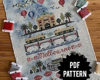 May include: A cross-stitch pattern featuring a colorful illustration of a city scene with buildings, trams, and the text "Melbourne, Australia". The pattern is printed on white paper and includes a black and white starburst with the text "PDF PATTERN".