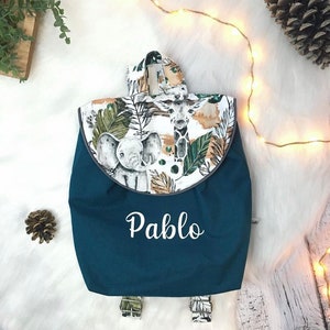 Op de afbeelding: Een donkerblauwe rugzak met een klep voorzien van een jungleprint met een olifant en een giraf op een witte achtergrond. De naam Pablo staat in witte letters op de voorkant van de tas. De rugzak heeft verstelbare schouderbanden.