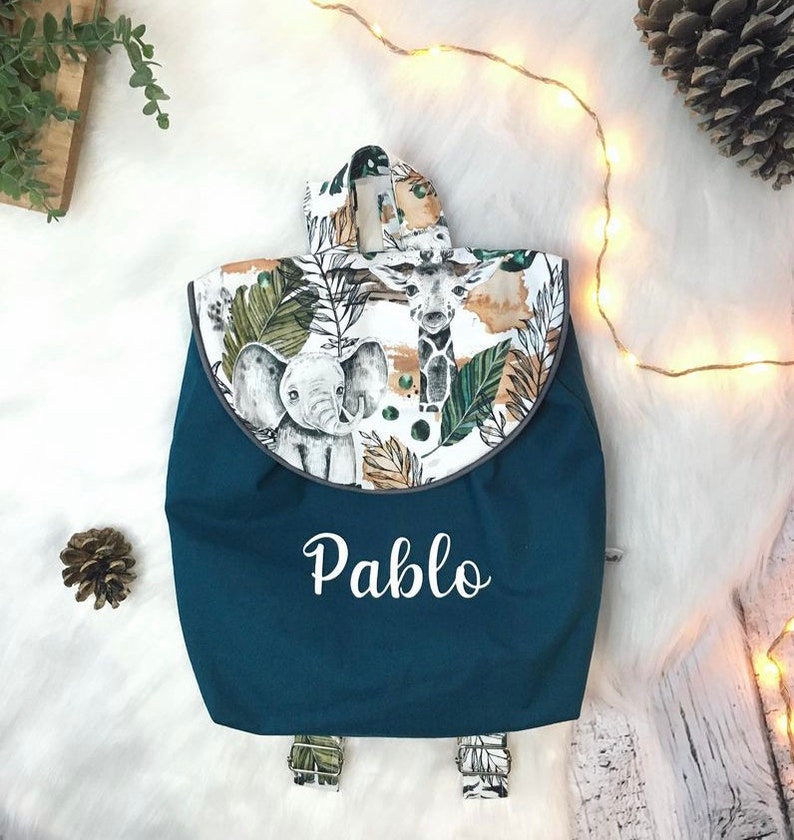 Op de afbeelding: Een donkerblauwe rugzak met een klep voorzien van een jungleprint met een olifant en een giraf op een witte achtergrond. De naam Pablo staat in witte letters op de voorkant van de tas. De rugzak heeft verstelbare schouderbanden.