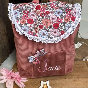 Op de afbeelding: Een roze corduroy rugzak met een bloemenprint flap en een witte kanten rand. De rugzak heeft een klein vakje met een bloemenstrik en de naam "Jade" geborduurd. Een roze bloem en stervormige lampjes staan op de voorgrond.