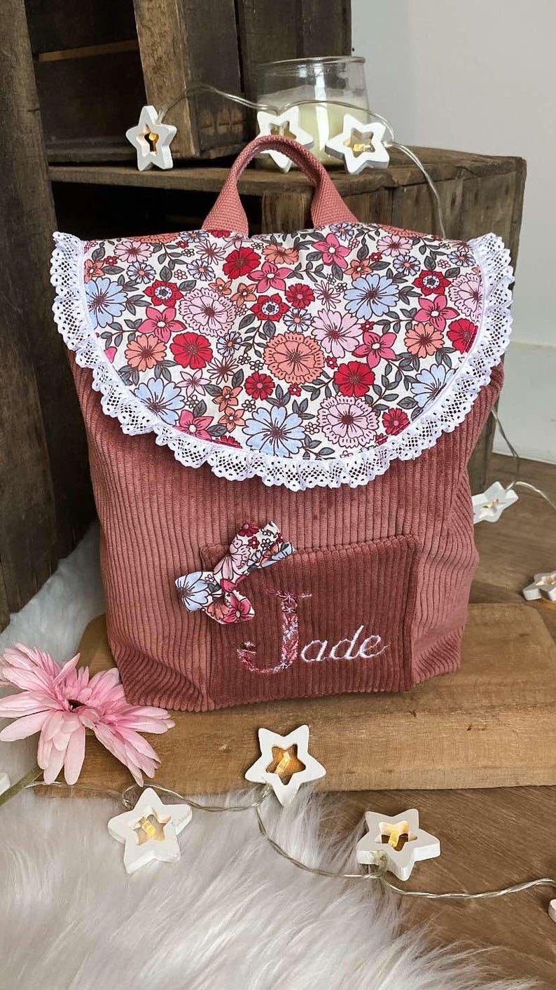 Op de afbeelding: Een roze corduroy rugzak met een bloemenprint flap en een witte kanten rand. De rugzak heeft een klein vakje met een bloemenstrik en de naam "Jade" geborduurd. Een roze bloem en stervormige lampjes staan op de voorgrond.