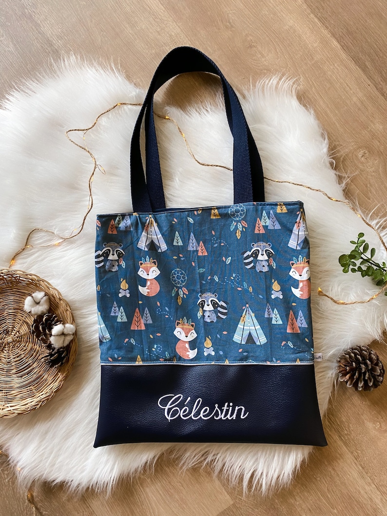 May include: A tote bag featuring a dark blue fabric top section with a repeating pattern of foxes, raccoons, teepees, and dreamcatchers. The bottom section is solid dark blue faux leather with the name C&eacute;lestin embroidered in white script.