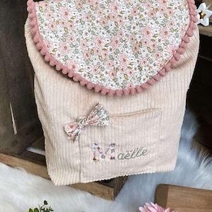 Op de afbeelding: Een lichtbeige corduroy rugzak met een bloemenprint flap en strik. De flap is afgezet met roze pompons. De naam "Ma&euml;lle" is geborduurd op de voorzak. De tas heeft een beige en wit gestreepte handvat.
