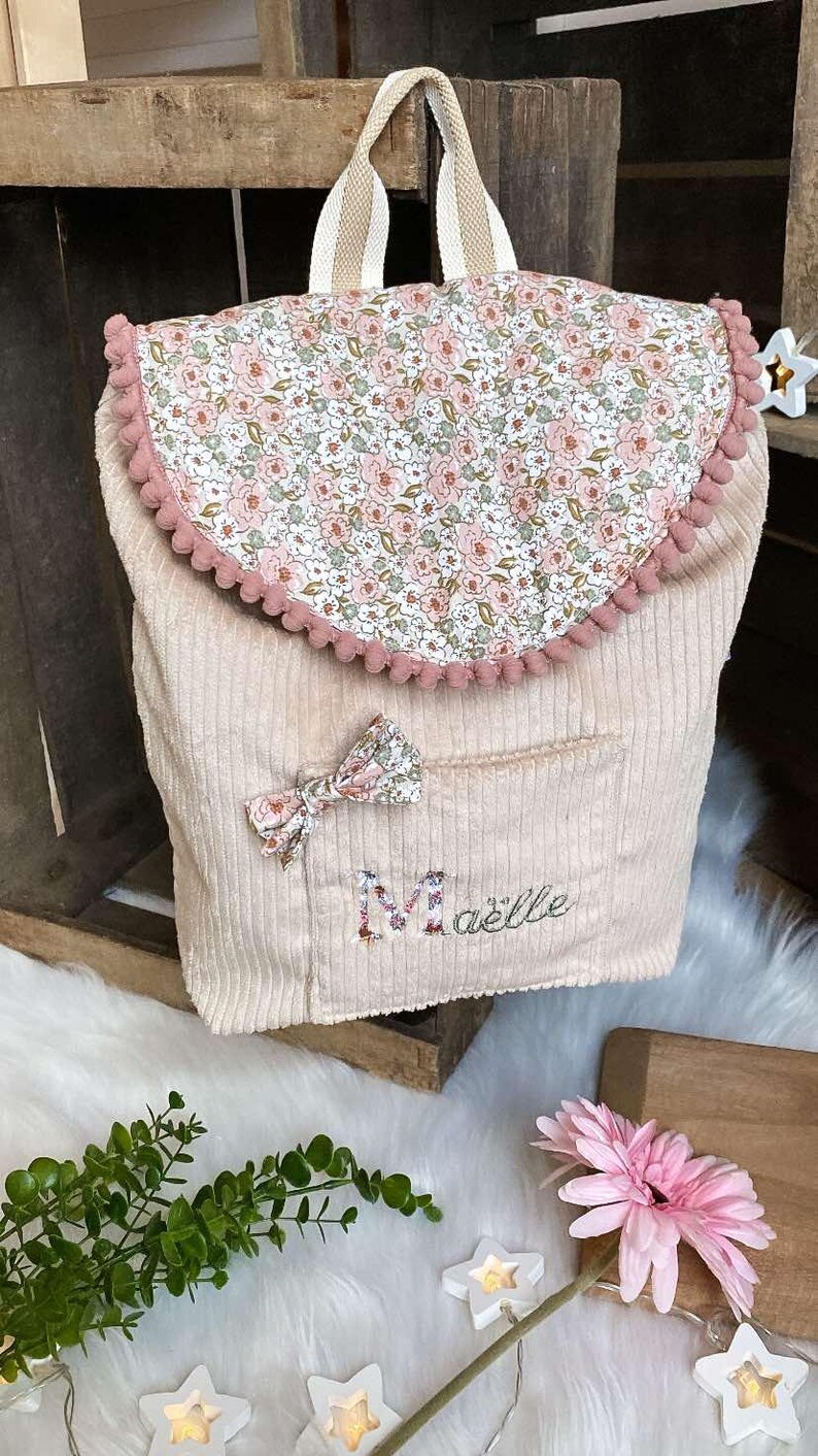Op de afbeelding: Een lichtbeige corduroy rugzak met een bloemenprint flap en strik. De flap is afgezet met roze pompons. De naam "Ma&euml;lle" is geborduurd op de voorzak. De tas heeft een beige en wit gestreepte handvat.
