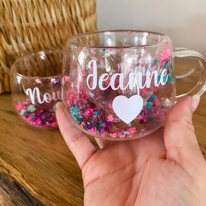 Op de afbeelding: Een heldere glazen mok gevuld met kleurrijke gedroogde bloemen en de naam "Jeanne" in witte letters, samen met een wit hart. Een kleiner glas met de naam "Now" is ook zichtbaar.