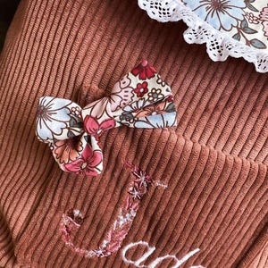Op de afbeelding: Close-up van een stoffige roze corduroy zak met een bloemenstrik en een bloemenpatroon. De naam "Jade" is geborduurd in witte letters. De strik heeft roze, rode en blauwe bloemen.