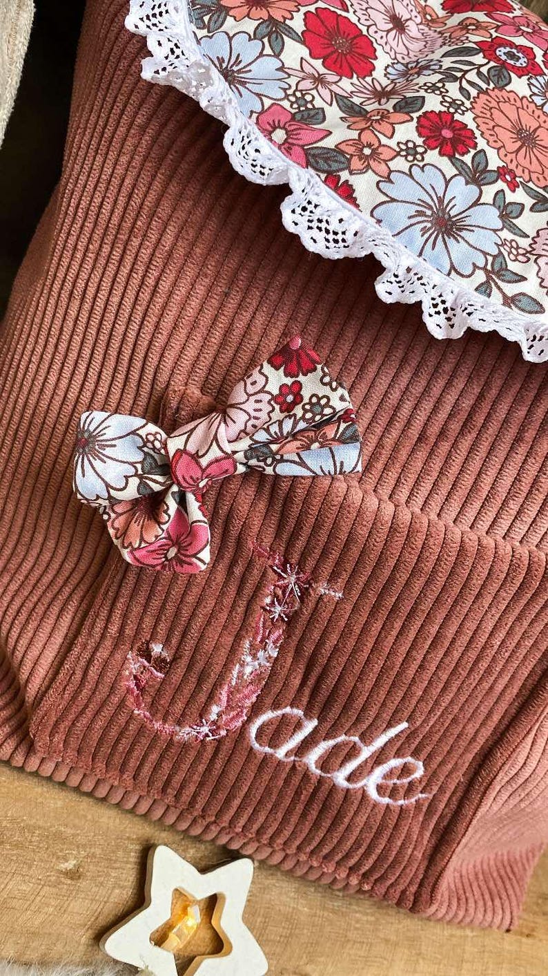 Op de afbeelding: Close-up van een stoffige roze corduroy zak met een bloemenstrik en een bloemenpatroon. De naam "Jade" is geborduurd in witte letters. De strik heeft roze, rode en blauwe bloemen.