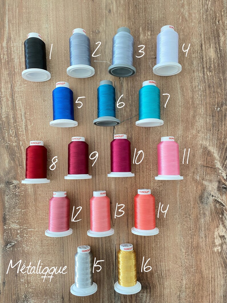 Op de afbeelding: Zestien klossen Gunold borduurgaren gerangschikt in vier rijen, genummerd van 1 tot 16. De kleuren omvatten zwart, diverse grijstinten, wit, blauw, teal, rood, magenta, roze, koraal, oranje, zilverkleurig en goudkleurig metallic garen.