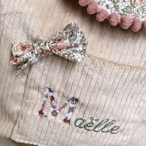 Op de afbeelding: Een beige corduroy item met een bloemenpatroon strik en afwerking. Het item heeft een bloemenpatroon zak en de naam "Ma&euml;llie" geborduurd in groen. Roze pompons omlijsten de bloemenstof.