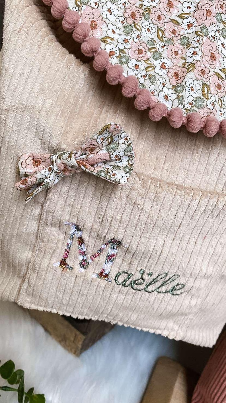 Op de afbeelding: Een beige corduroy item met een bloemenpatroon strik en afwerking. Het item heeft een bloemenpatroon zak en de naam "Ma&euml;llie" geborduurd in groen. Roze pompons omlijsten de bloemenstof.