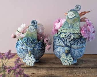 以下が含まれることがあります： ピンクの花で満たされた、2つの鳥の形をしたセラミック花瓶。花瓶は青色で、複雑な花の模様と「LOVE」と書かれた装飾的なペンダントが付いています。鳥はティールと青色のディテールを持っています。