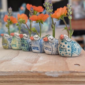 May include: Six ceramic bird-shaped vases in a row, each with a different blue and white pattern and the word "LOVE". The vases hold orange flowers and purple lavender sprigs. The arrangement sits on a wooden surface.