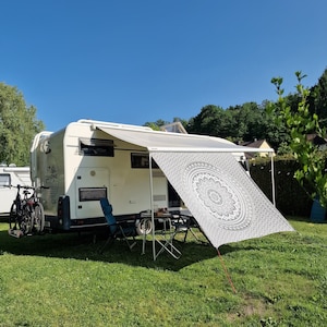 May include: A white camper van with a white awning and a black and white mandala patterned canopy. The camper van has a sign that says "Camping Vibes" on the side. There are two chairs and a table set up in front of the camper van.