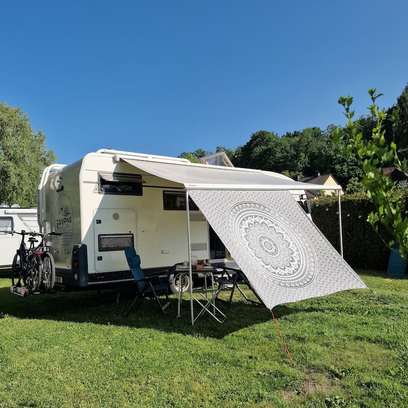May include: A white camper van with a white awning and a black and white mandala patterned canopy. The camper van has a sign that says "Camping Vibes" on the side. There are two chairs and a table set up in front of the camper van.