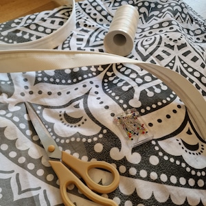 May include: A white fabric with a black and white patterned design. There is a white fabric trim, a spool of white thread, a box of colorful sewing pins, and a pair of yellow-handled scissors on the fabric.