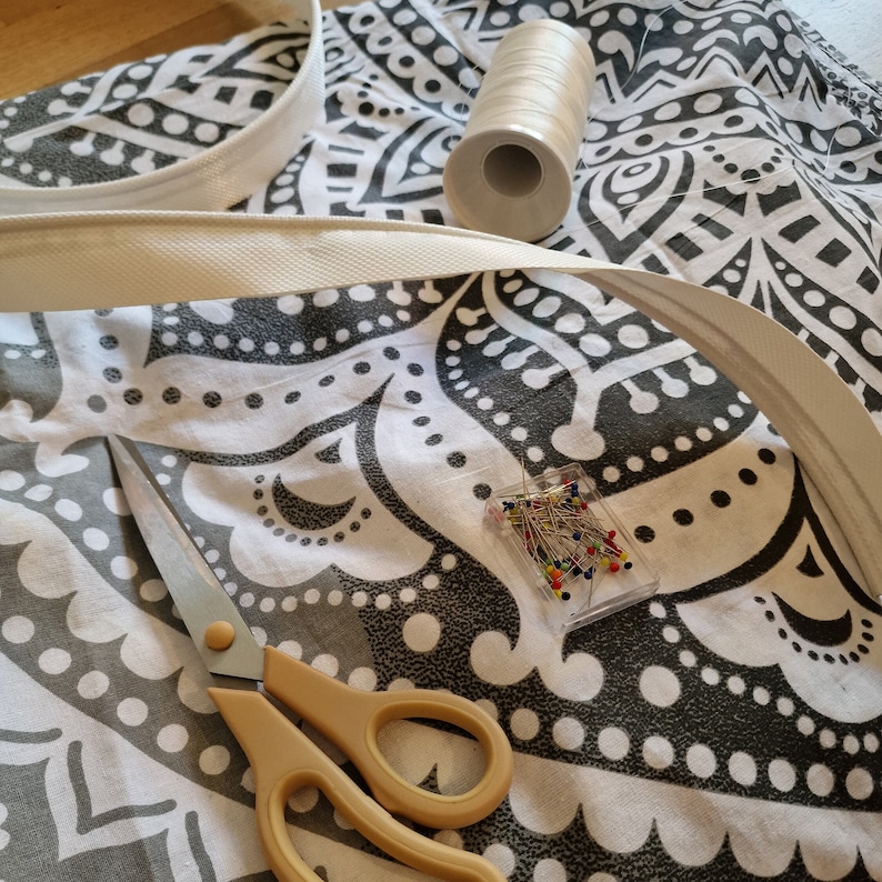 May include: A white fabric with a black and white patterned design. There is a white fabric trim, a spool of white thread, a box of colorful sewing pins, and a pair of yellow-handled scissors on the fabric.
