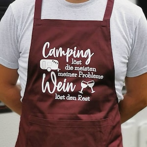 Könnte beinhalten: Eine dunkelrote Schürze mit einem weißen Aufdruck, der "Camping löst die meisten meiner Probleme Wein löst den Rest" lautet, mit einem Bild eines Wohnwagens und zwei Weingläsern.