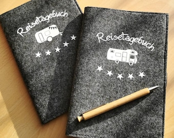 Puede incluir: Dos cuadernos cubiertos de fieltro gris con el texto "Reisetagebuch" y un gráfico de autocaravana blanco. El cuaderno de la derecha tiene un bolígrafo de madera en la parte superior.
