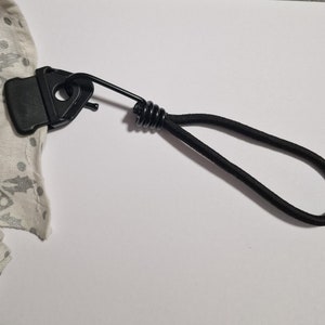May include: A black plastic clip with a spring and a black elastic cord attached. The clip is attached to a white fabric with a pattern.
