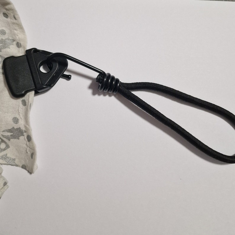 May include: A black plastic clip with a spring and a black elastic cord attached. The clip is attached to a white fabric with a pattern.