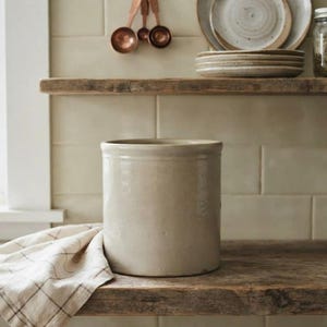 Può includere: Un contenitore cilindrico in ceramica color beige chiaro con una finitura liscia e opaca. Il recipiente presenta un design semplice con pareti dritte ed è posizionato accanto a un panno a quadri bianchi e marroni su una superficie in legno.