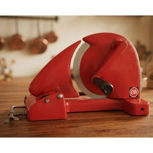 May include: A vintage red manual food slicer featuring a circular serrated metal blade, a hand crank with a black handle, and a metal clamp mechanism for securing the device to a surface. The brand name EVA is printed on a small white label.