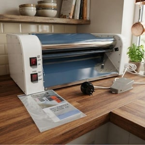 May include: A white and blue electric dough sheeter machine featuring two black power switches on the side panel labeled ON and OFF. The device includes a foot pedal controller, a power cord, and a clear plastic bag containing an instruction manual.