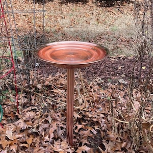 Pode incluir: Um banho para pássaros na cor cobre com uma bacia circular larga de superfície lisa e reflexiva, montada sobre um pedestal central. O acabamento metálico exibe tons quentes de castanho-avermelhado e uma estrutura aberta.