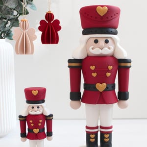 Könnte beinhalten: Zwei Nussknacker-Figuren in unterschiedlichen Größen, mit einer dunkelroten Uniform, schwarzen Akzenten, weißen Hosen und goldenen herzförmigen Knöpfen. Jeder Nussknacker hat einen weißen Schnurrbart, einen hohen roten Hut und einen weißen Sockel.