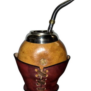 Puede incluir: Un mate de calabaza redondo de color beige con textura natural lisa y un borde de metal plateado pulido. La taza está envuelta en una funda de cuero rojo oscuro con un cordón decorativo en la parte frontal e incluye una bombilla de metal larga y curva.