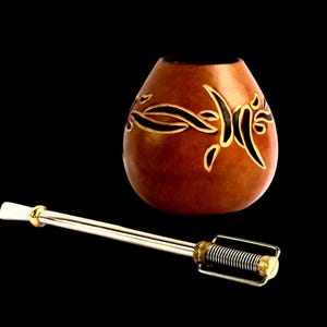 Puede incluir: Una calabaza marrón con un diseño negro y dorado, utilizada para beber mate, con una bombilla de paja de plata.