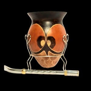 Può includere: Una tazza di mate a forma di zucca con un supporto e una cannuccia in metallo argentato. La tazza è marrone con segni arancioni e neri a forma di gufo. La cannuccia ha una finitura argentata con dettagli dorati.
