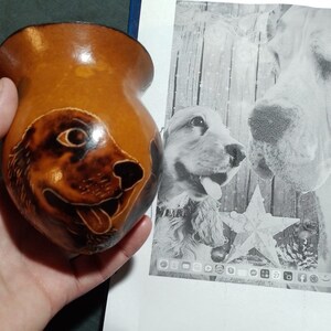 Puede incluir: Una calabaza marrón con una imagen tallada de la cara de un perro. La calabaza está siendo sostenida en la mano de una persona. El fondo es una imagen borrosa de dos perros.