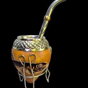 Può includere: Una tazza di mate a forma di zucca con bordo e supporto argentati, con un'incisione di pesce. La tazza include una cannuccia bombilla argentata. Lo sfondo è nero.