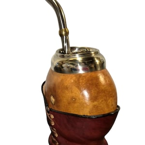 Puede incluir: Un recipiente para mate hecho de calabaza con superficie color canela, borde de metal plateado pulido y una bombilla de metal plateado a juego con una banda dorada. La base está recubierta por una funda de cuero marrón oscuro con cordones.