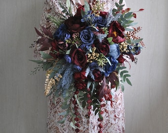 May include: A cascading bridal bouquet with deep red and blue flowers, accented with gold and greenery. The bouquet features a long trailing cascade of red berries.