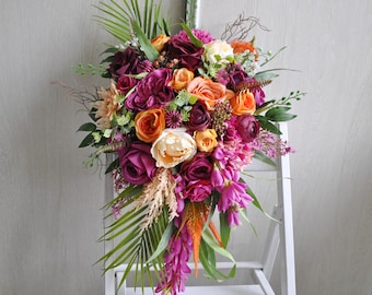 May include: A cascading floral arrangement featuring a variety of flowers in shades of purple, orange, and cream. The bouquet includes roses, peonies, and other blooms, accented with green foliage and palm leaves. The arrangement is displayed on a white stand.